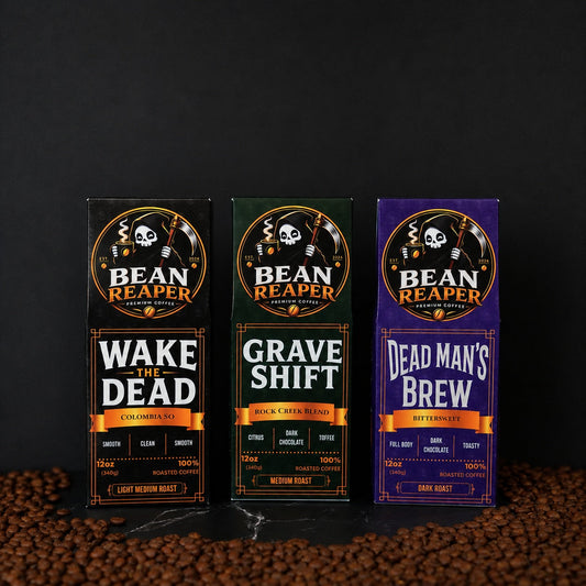 Three Bean Reaper 12 oz coffee bags side by side — Wake The Dead Colombia single origin, Grave Shift medium roast blend and Dead Man's Brew Brazilian dark roast — standing on whole coffee beans